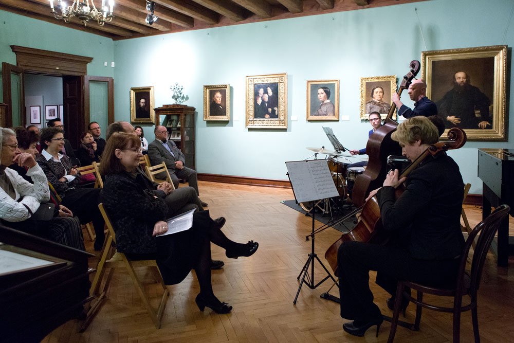 Trio muzyków — wiolonczela, kontrabas i perkusja — występuje w sali z zielonymi ścianami ozdobionymi złotymi ramami portretów, a publiczność siedzi na składanych krzesłach; widoczna drewniana podłoga, belkowany sufit i żyrandol.