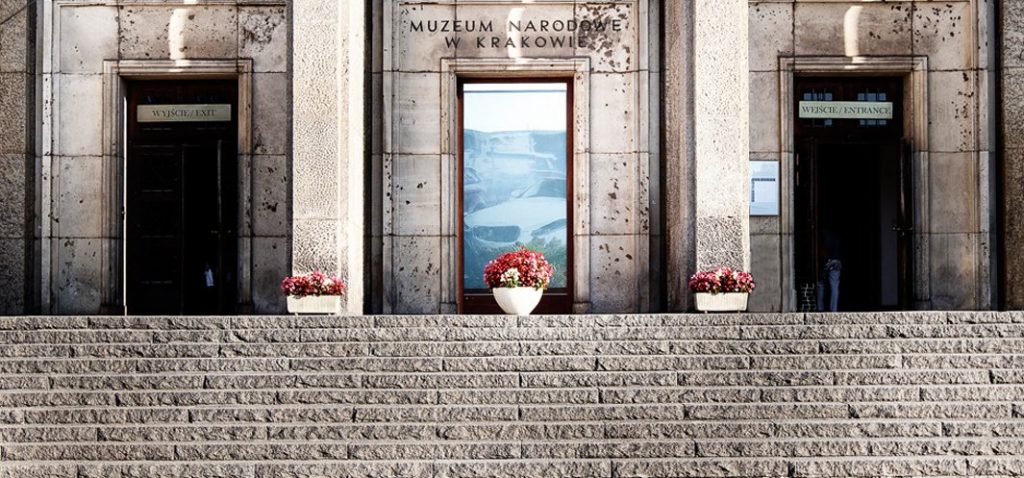 Kamienna fasada Muzeum Narodowego w Krakowie z szerokimi stopniami prowadzącymi do trzech prostokątnych wejść — na środku pod napisem "MUZEUM NARODOWE W KRAKOWIE" przeszklone drzwi z dużym plakatem i białą donicą z czerwonymi kwiatami, po bokach dwa wejścia z tabliczkami "WYJŚCIE/EXIT" i "WEJŚCIE/ENTRANCE" oraz mniejsze donice z czerwonymi kwiatami; w prawej wnęce widoczna dolna część ciała osoby.