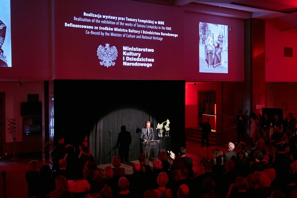 Widok sali wypełnionej siedzącą publicznością podczas przemówienia mężczyzny przy mównicy na niewielkiej scenie oświetlonej reflektorem, z czerwonym oświetleniem bocznym; na ścianie nad sceną duży ekran z tekstem po polsku i angielsku informującym o wystawie Tamary Łempickiej oraz herbem i napisem Ministerstwo Kultury i Dziedzictwa Narodowego, dwoma projekcjami zdjęć po bokach i wazonem z białymi kwiatami przy mównicy.