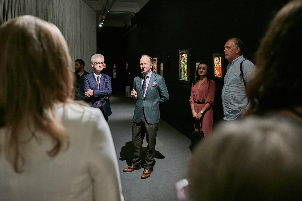 Grupa osób stoi w ciemnej sali wystawowej przy oświetlonych obrazach; na środku stoi mężczyzna w szarym garniturze gestykulujący, obok po lewej mężczyzna w granatowym garniturze z założonymi rękami, po prawej kobieta w różowej sukience trzymająca torebkę i mężczyzna w jasnej koszuli z plecakiem, a w pierwszym planie widać rozmyte sylwetki widzów.