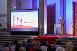 Publiczność siedzi w sali i ogląda osobę przemawiającą przy przezroczystej mównicy po prawej stronie czerwonej sceny, obok dużego ekranu projekcyjnego z czerwoną grafiką i widocznym tekstem, we wnętrzu z wysokimi kolumnami i fioletowo-niebieskim oświetleniem.