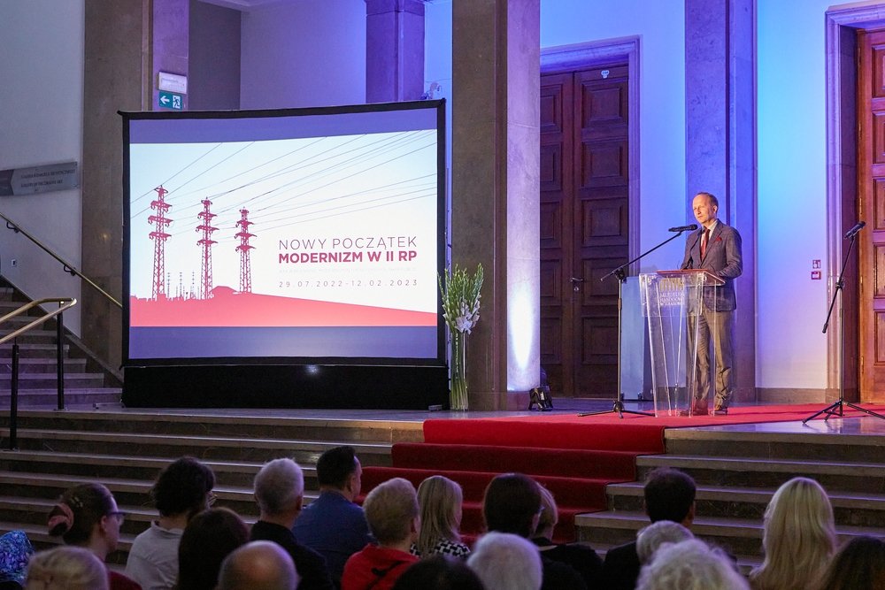 Mężczyzna stoi przy przezroczystym pulpicie na scenie z czerwonym dywanem i przemawia do publiczności, obok duży ekran projekcyjny wyświetlający planszę „Nowy początek Modernizm w II RP” z ilustracją słupów energetycznych, a przed sceną siedzą widzowie, w tle kolumny i drewniane drzwi.