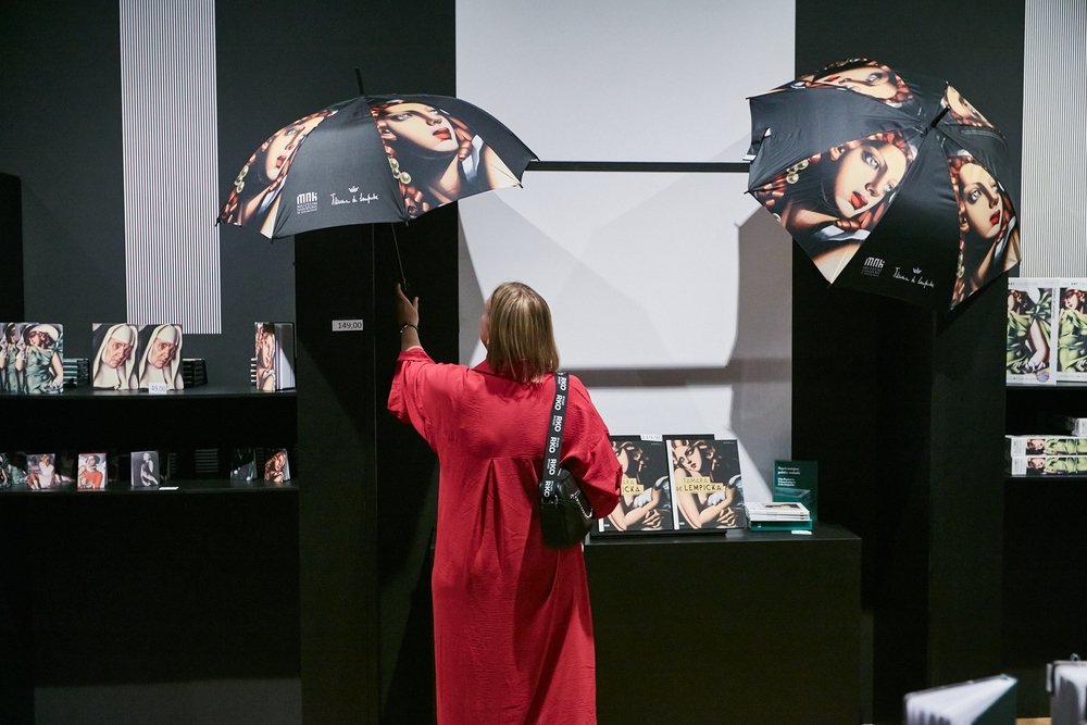 Osoba w czerwonej sukni widziana od tyłu trzyma czarną parasolkę z nadrukiem portretu i stoi przed czarnym stoiskem z drugą podobną parasolką oraz półkami z książkami i reprodukcjami obrazów.
