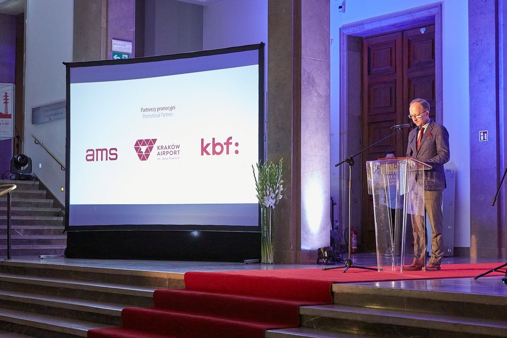 Wnętrze sali z podwyższoną sceną i czerwonym dywanem, po lewej duży ekran projekcyjny wyświetlający logotypy "ams", "Kraków Airport" i "kbf:", po prawej mężczyzna w garniturze i okularach stoi przy przezroczystym pulpicie i mikrofonie, obok wysoki wazon z białymi kwiatami, w tle marmurowe kolumny i drewniane podwójne drzwi oświetlone na niebiesko.