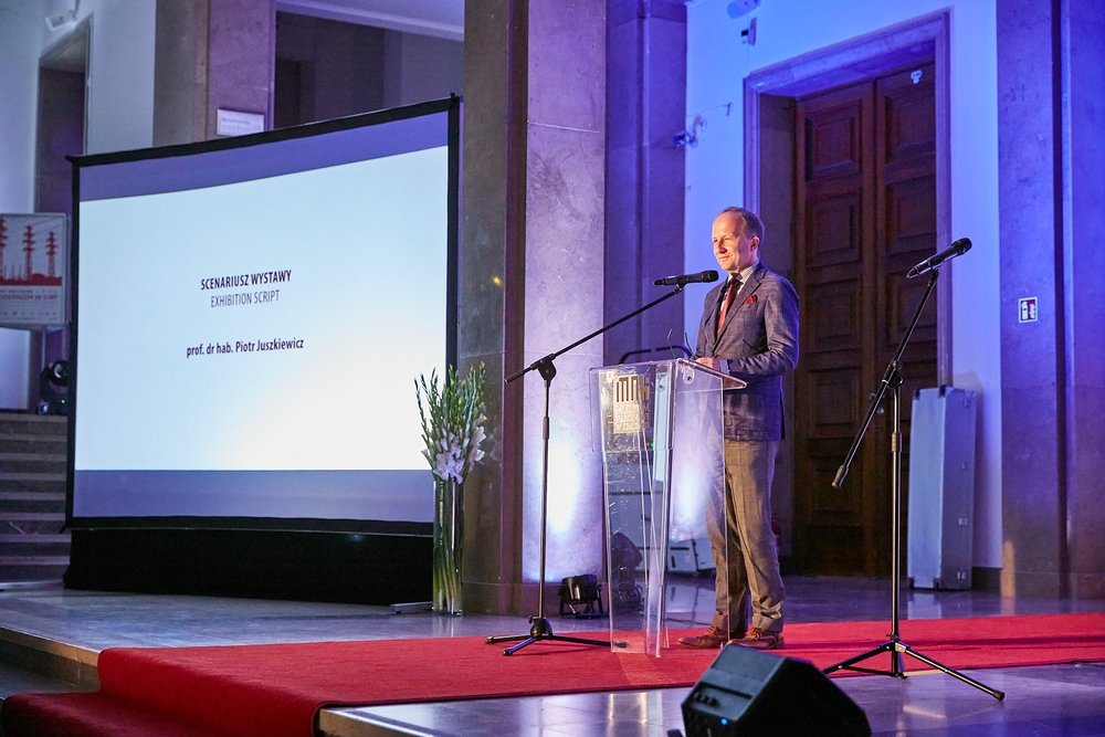 Mężczyzna w garniturze stoi za przezroczystą mównicą na czerwonym dywanie z dwoma mikrofonami, po lewej duży ekran projekcyjny z napisem „SCENARIUSZ WYSTAWY / EXHIBITION SCRIPT” i nazwiskiem „prof. dr hab. Piotr Juszkiewicz”, przy ekranie wysoki wazon z białymi kwiatami, w tle marmurowe kolumny i drewniane drzwi oświetlone na niebiesko.