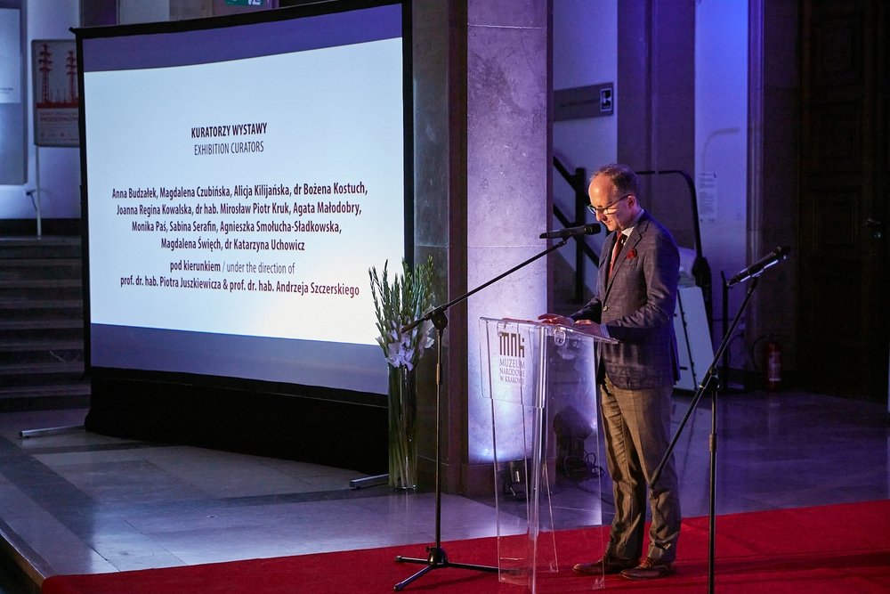 Mężczyzna w marynarce stoi przy przezroczystym podium z mikrofonem i widocznym napisem muzeum, trzyma kartkę, obok duży ekran projekcyjny wyświetlający tytuł "KURATORZY WYSTAWY" i listę nazwisk, na podłodze czerwony dywan, widoczna wysoka kolumna i wazon z białymi kwiatami.