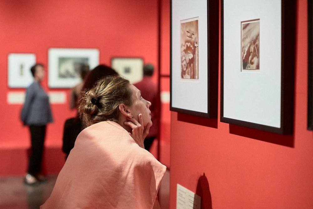 Kobieta z upiętymi włosami, opierająca dłoń o brodę, pochylona nad dwoma oprawionymi zdjęciami na czerwonej ścianie galerii, w tle rozmyte postacie i inne prace.