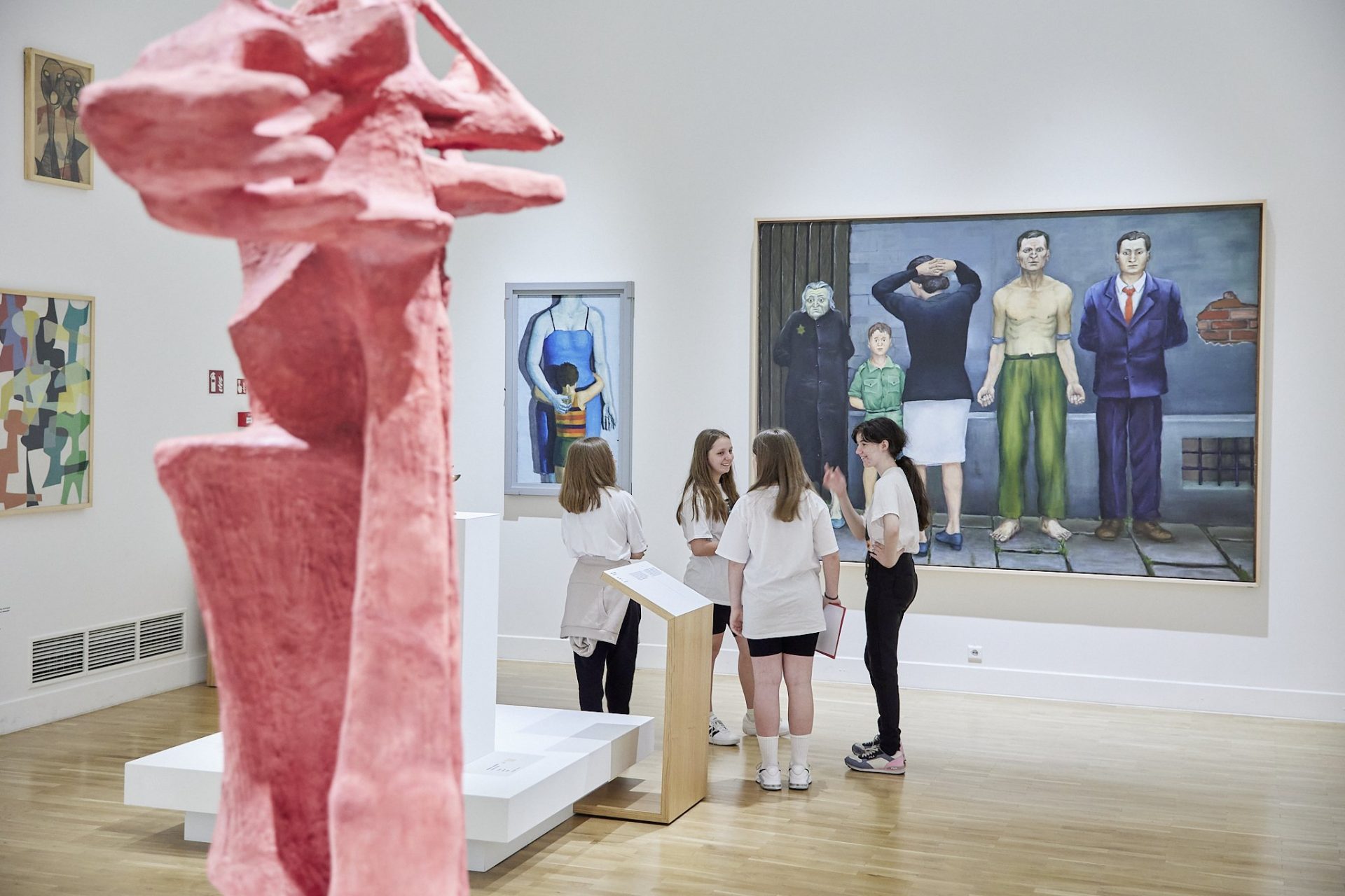 Galeria sztuki z białymi ścianami i drewnianą podłogą, z dużą różową abstrakcyjną rzeźbą na pierwszym planie po lewej, czwórką młodych osób w jasnych koszulkach stojących przy drewnianym pulpicie w środku oraz wielkoformatowym obrazem po prawej przedstawiającym kilka stojących postaci i dodatkowymi mniejszymi obrazami na ścianach.