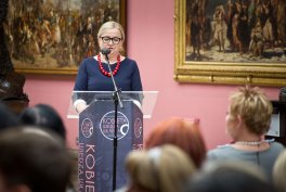Kobieta w okularach w ciemnej sukience z białym kołnierzykiem i czerwonym naszyjnikiem stoi za przezroczystą mównicą i przemawia do siedzącej publiczności, na różowej ścianie za nią wiszą dwa duże klasyczne obrazy w złotych ramach.