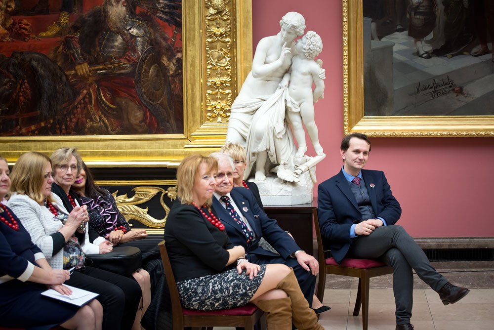 Grupa siedzących osób w formalnych ubraniach na krzesłach w sali galerii z różową ścianą, na pierwszym planie kobiety (niektóre w czerwonych koralach) i mężczyźni, za nimi duża biała marmurowa rzeźba kobiety z dzieckiem oraz bogato złocone ramy wielkich obrazów.