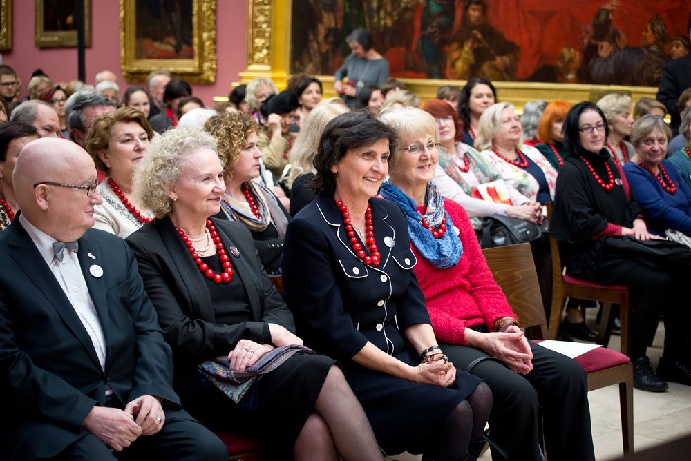 Publiczność siedząca w rzędach w sali z dużymi, złoconymi ramami obrazów — przede wszystkim kobiety w eleganckich strojach, wiele nosi charakterystyczne czerwone korale, na pierwszym planie kilka osób siedzi obok siebie na drewnianych krzesłach.