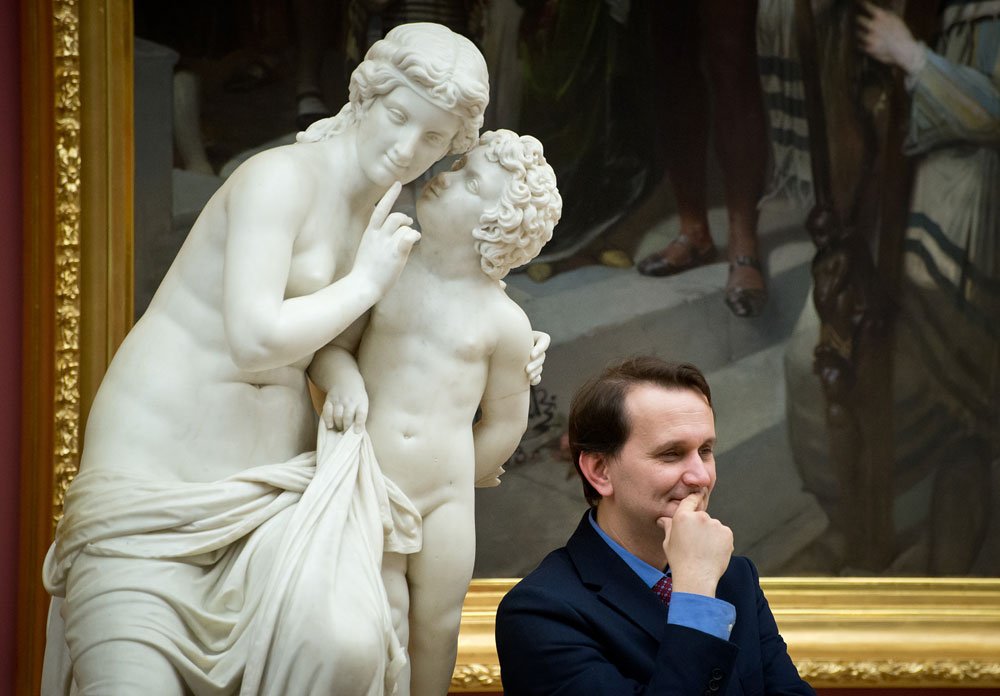 Biała marmurowa rzeźba kobiety przytulającej dziecko, kobieta przykłada palec do ust, obok siedzi mężczyzna w garniturze z ręką przy brodzie, a w tle widoczny duży obraz w złotej ramie.