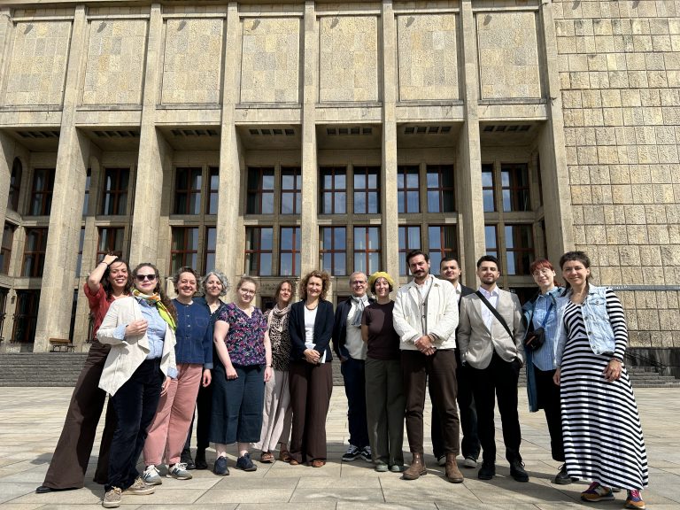 Grupowe zdjęcie około czternastu dorosłych stojących w jednym rzędzie przed masywną kamienną fasadą budynku z wysokimi pionowymi filarami, wieloma prostokątnymi oknami i stopniami u podstawy.