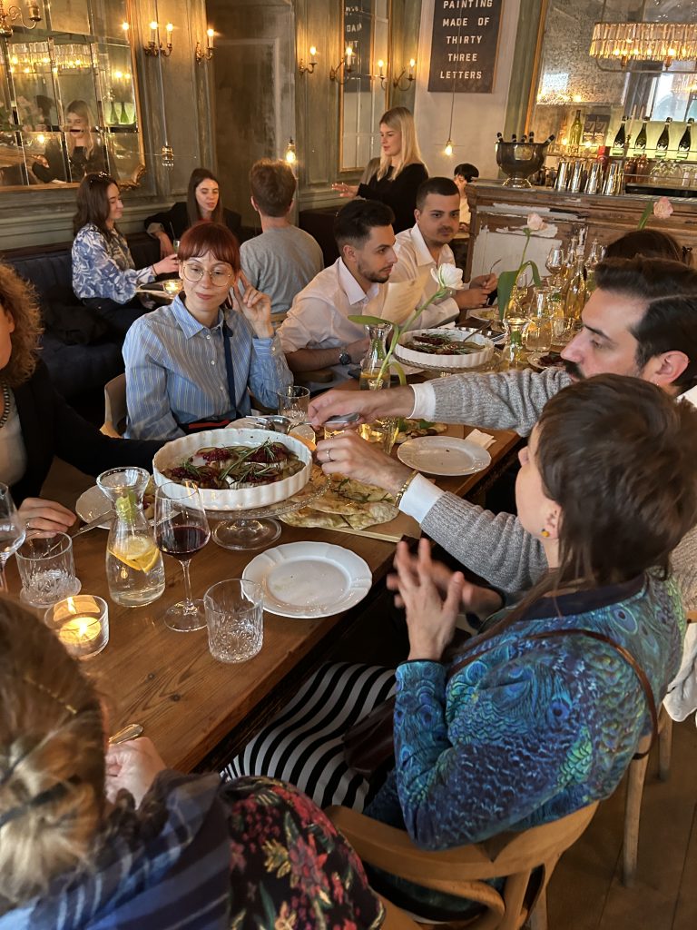 Kilka osób siedzi przy długim drewnianym stole w przytulnie oświetlonej restauracji; na stole są misy z jedzeniem, talerze, kieliszki i karafka z plasterkiem cytryny, zapalona świeca oraz wąskie wazoniki z białymi kwiatami, a w tle widać lustrzaną ścianę, kinkiety i bar z butelkami.
