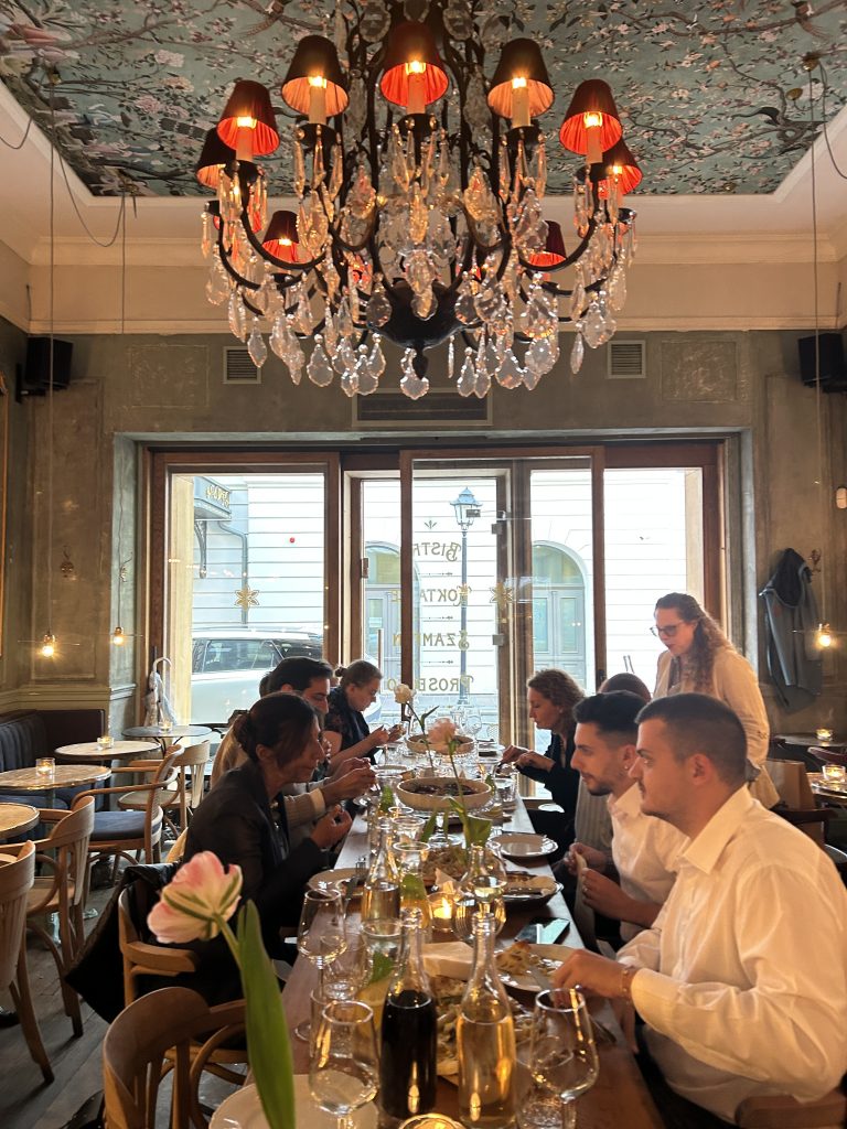 Wnętrze restauracji z dekoracyjnym sufitem w kwiatowy wzór i dużym kryształowym żyrandolem z czerwonymi kloszami, długi drewniany stół zastawiony talerzami, kieliszkami i butelkami, kilkoma osobami jedzącymi przy stole, wysokimi drzwiami i oknami wychodzącymi na ulicę oraz w pierwszym planie pąkiem różowego tulipana.