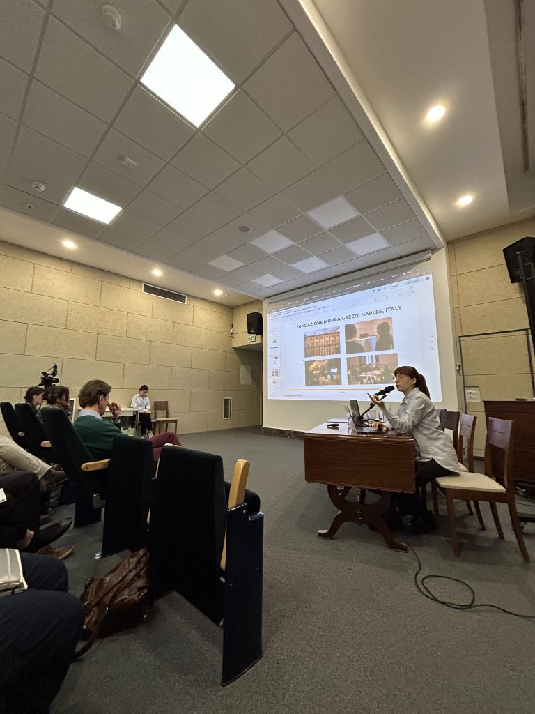 Pomieszczenie konferencyjne z sufitowymi panelami i punktowymi światłami; po prawej siedzi osoba przy drewnianym stole z mikrofonem i laptopem, za nią na dużym ekranie wyświetlona prezentacja z miniaturami zdjęć i napisem "FONDAZIONE MORRA GRECO, NAPLES, ITALY"; po lewej rzędy foteli z kilkoma uczestnikami i kamerą, na podłodze widoczny zwinięty kabel.
