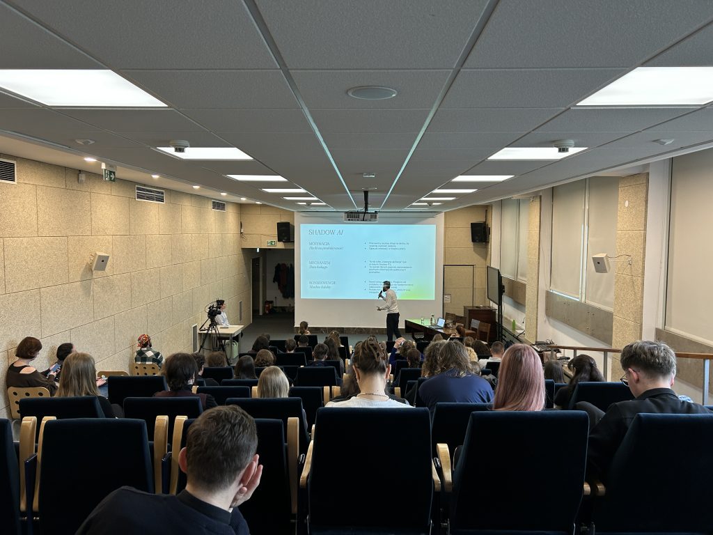 Zdjęcie wnętrza wykładowej sali widzianej z tyłu: rzędy ciemnych foteli z siedzącą publicznością, centralny ekran projekcyjny z wyświetlonym slajdem, stojący prowadzący obok ekranu, kamera na statywie po lewej, jasne sufitowe panele z oświetleniem oraz beżowe ściany z głośnikami i kratkami wentylacyjnymi.