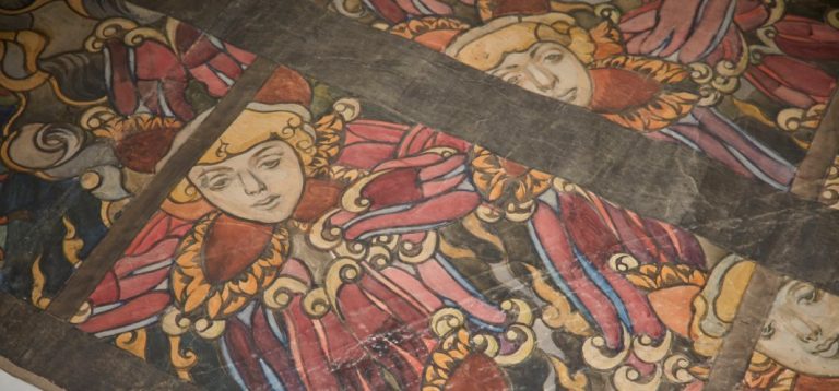 Fragment malowanej dekoracji przedstawiający powtarzające się stylizowane twarze w złoto-czerwonych, falbaniastych strojach z ozdobnymi zawijasami, rozmieszczone w polach oddzielonych ciemnymi drewnianymi belkami.