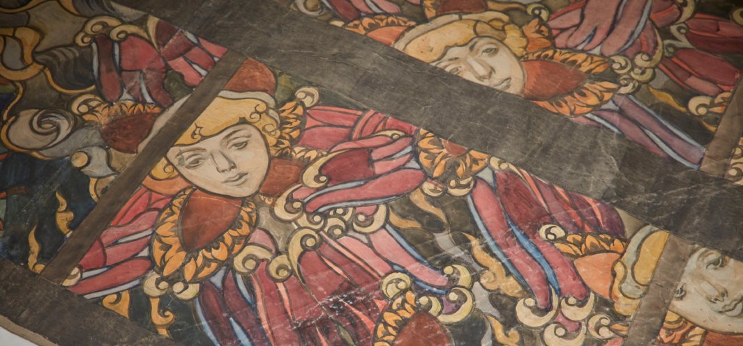 Fragment malowanej dekoracji przedstawiający powtarzające się stylizowane twarze w złoto-czerwonych, falbaniastych strojach z ozdobnymi zawijasami, rozmieszczone w polach oddzielonych ciemnymi drewnianymi belkami.