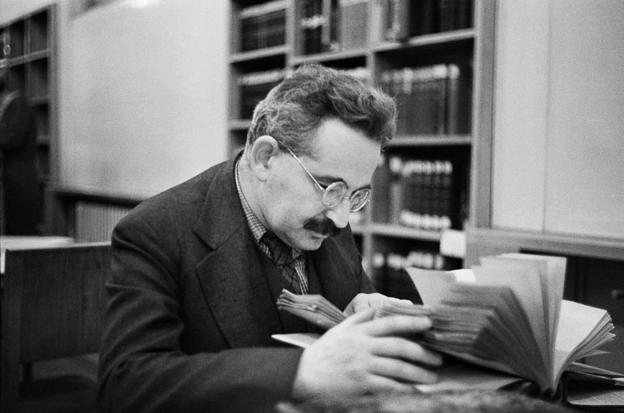 Czarno-białe zdjęcie mężczyzny w okularach i wąsami w marynarce siedzącego przy stoliku w bibliotece i przeglądającego otwartą książkę, za nim regały z książkami.