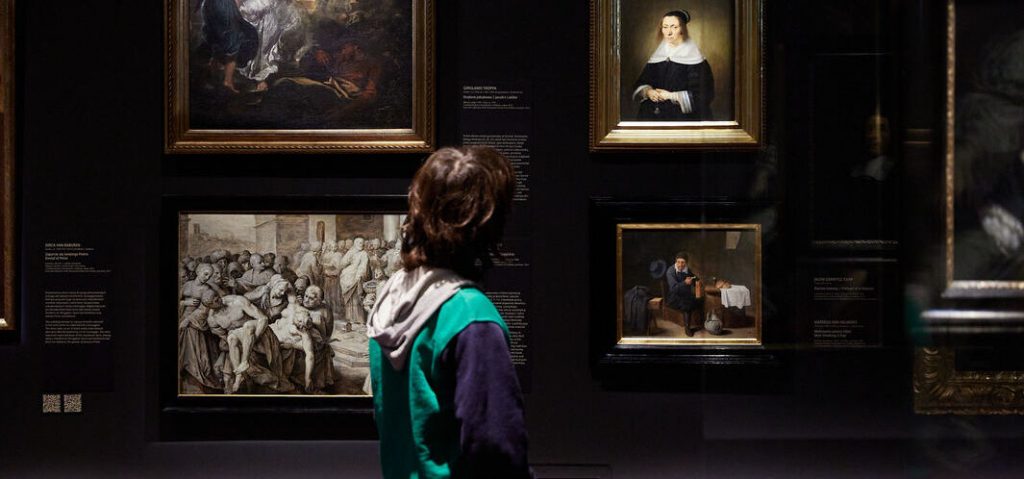 Osoba widziana od tyłu w zielono‑szarej bluzie stoi przed ciemną ścianą galerii, na której wiszą oprawione dzieła: u góry ciemny obraz olejny, poniżej po lewej duża monochromatyczna rycina przedstawiająca grupę postaci, po prawej portret kobiety w białym kołnierzu oraz mniejszy obraz wnętrza z siedzącym mężczyzną, a obok widoczne są tabliczki informacyjne i odbicia na szklanych ramach.