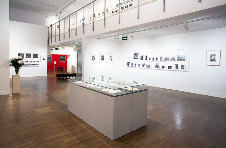 Wnętrze jasnej galerii wystawowej z białymi ścianami i drewnianą podłogą; centralnie ustawiona szklana witryna na białym podeście, na ścianach oprawione fotografie i układane kolaże, balkon z metalową balustradą z dodatkowymi zdjęciami, czerwony fragment ściany w tle, ławka wypoczynkowa i wazon z białymi kwiatami po lewej oraz oświetlenie sufitowe.