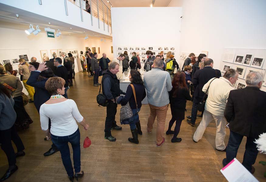 Wnętrze jasnej galerii sztuki z wieloma osobami oglądającymi czarno‑białe zdjęcia eksponowane na białych ścianach i na centralnym stoliku, z drewnianą podłogą, sufitowymi reflektorami i balkonem z poręczą na piętrze.