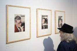 Trzy pionowe, oprawione portrety w jasnych drewnianych ramkach zawieszone obok siebie na białej ścianie galerii; po prawej stoi osoba o długich siwych włosach w czarnym kapeluszu i ciemnym płaszczu, oglądająca zdjęcia, a jej cień pada na ścianę; pod lewą ramką widoczna mała tabliczka z opisem.