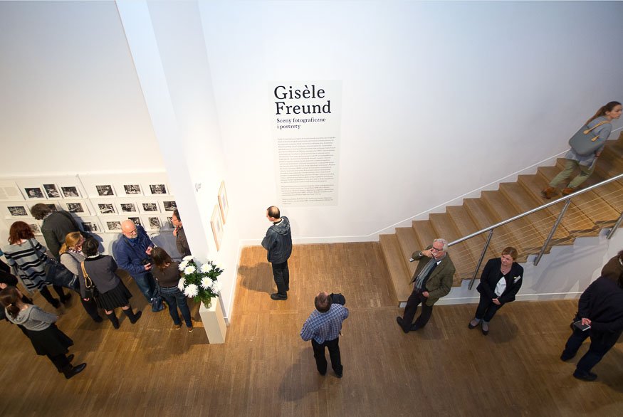 Widok z góry na jasne wnętrze galerii: duża biała ściana z tytułem wystawy "Gisèle Freund" i tekstem, seria małych czarno-białych fotografii na lewej ścianie oglądana przez grupę ludzi, postument z bukietem białych kwiatów przy słupie, kilka osób stojących na drewnianej podłodze oraz schody z prawej strony z metalową balustradą.