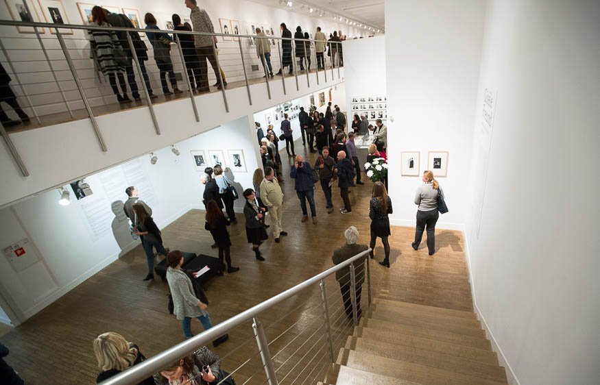 Widok wnętrza galerii z górnych schodów na dwupoziomową, białą przestrzeń wystawową z drewnianą podłogą, metalową balustradą i punktowym oświetleniem, gdzie liczne osoby na obu poziomach oglądają oprawione zdjęcia i prace wiszące na ścianach.